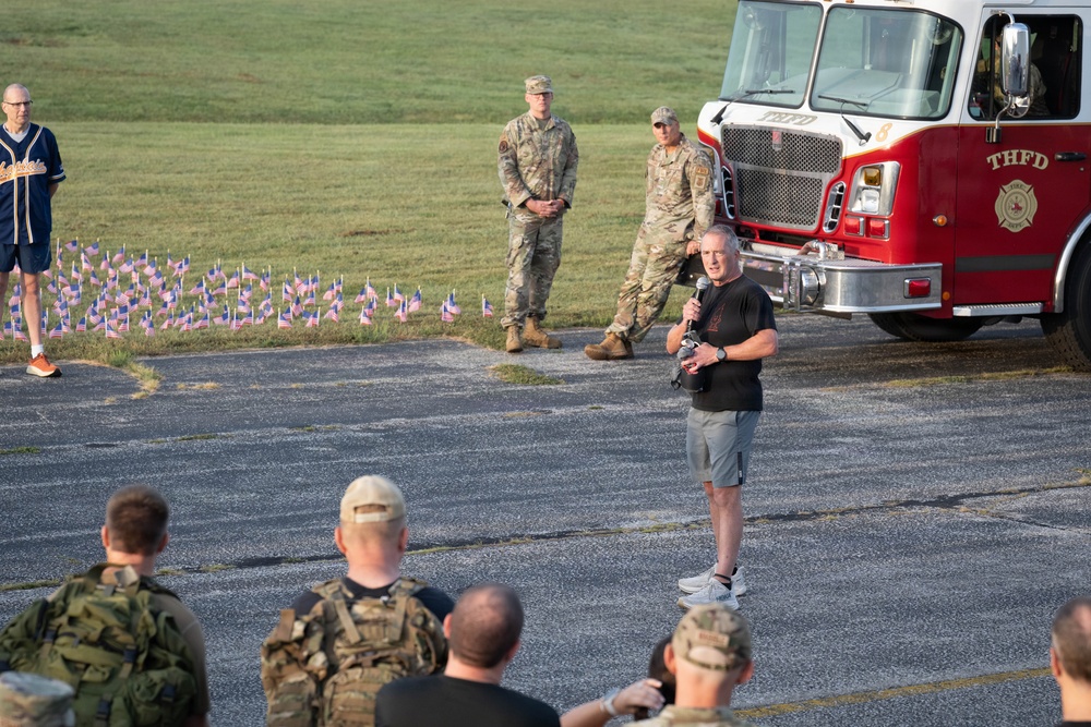 An outdoor gathering appears to be a military or memorial event, likely a 9/11 commemoration based on the small American flags arranged in the grass and the presence of a fire truck marked 'THFD'. A civilian man in a black t-shirt and shorts is speaking into a microphone while military personnel in camouflage uniforms listen. The event takes place on a paved area adjacent to a grassy field during daylight hours.