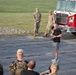 An outdoor gathering appears to be a military or memorial event, likely a 9/11 commemoration based on the small American flags arranged in the grass and the presence of a fire truck marked 'THFD'. A civilian man in a black t-shirt and shorts is speaking into a microphone while military personnel in camouflage uniforms listen. The event takes place on a paved area adjacent to a grassy field during daylight hours.