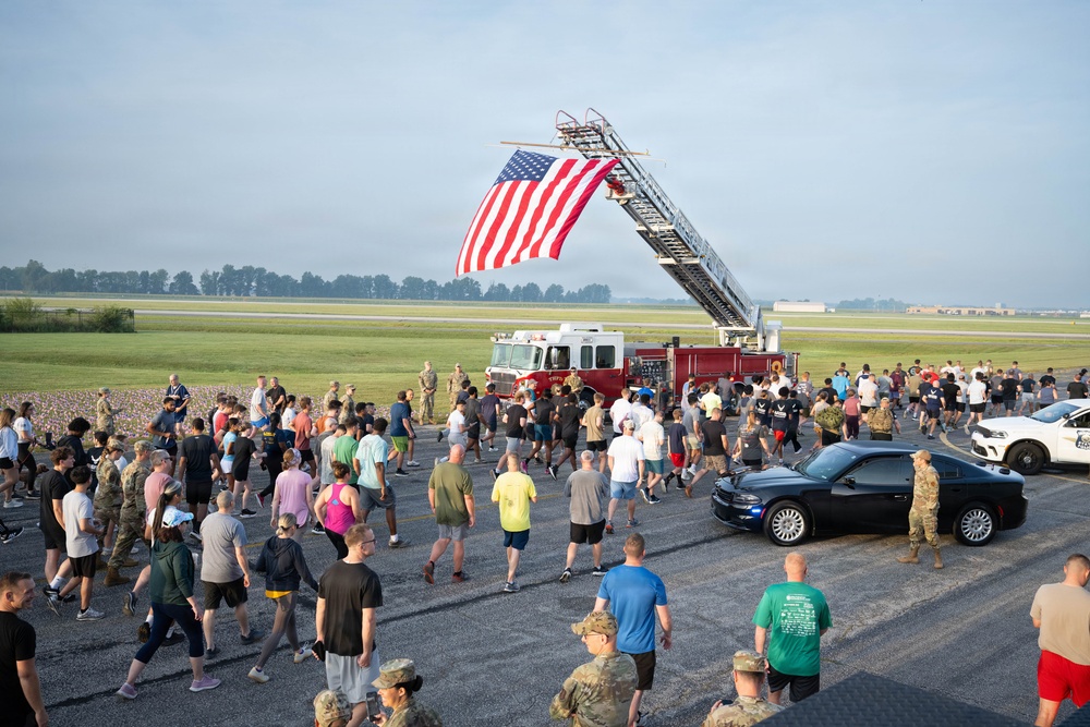 A large group of military personnel and civilians participate in what appears to be a memorial run or fitness event on a military airfield tarmac. A prominent American flag is displayed hanging from an extended fire truck ladder, serving as a ceremonial focal point for the gathering. The event appears to be a 9/11 memorial run or similar patriotic commemorative fitness event based on the setting and flag display.