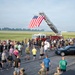 A large group of military personnel and civilians participate in what appears to be a memorial run or fitness event on a military airfield tarmac. A prominent American flag is displayed hanging from an extended fire truck ladder, serving as a ceremonial focal point for the gathering. The event appears to be a 9/11 memorial run or similar patriotic commemorative fitness event based on the setting and flag display.