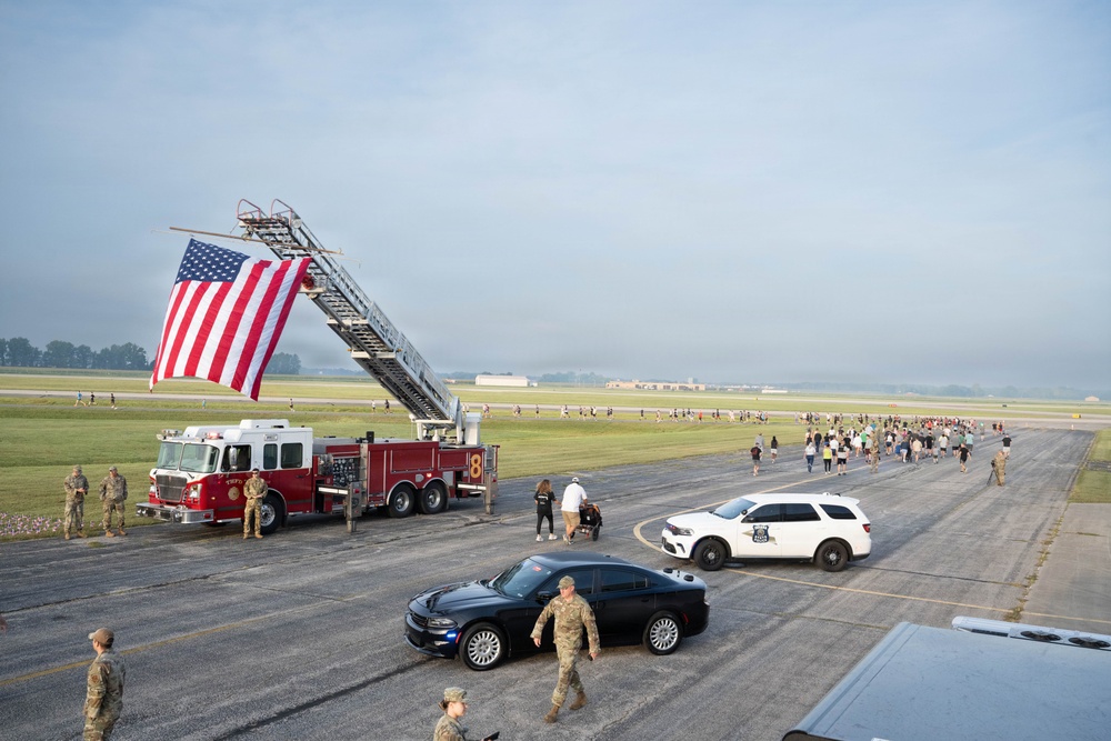 An outdoor event on a military airfield tarmac featuring a large American flag suspended from a fire truck's extended aerial ladder. Military personnel in camouflage uniforms are stationed along the route while a large group of civilians and participants walk or run along the runway. The scene appears to be a commemorative run or walk event, likely on a military installation.