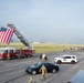 An outdoor event on a military airfield tarmac featuring a large American flag suspended from a fire truck's extended aerial ladder. Military personnel in camouflage uniforms are stationed along the route while a large group of civilians and participants walk or run along the runway. The scene appears to be a commemorative run or walk event, likely on a military installation.