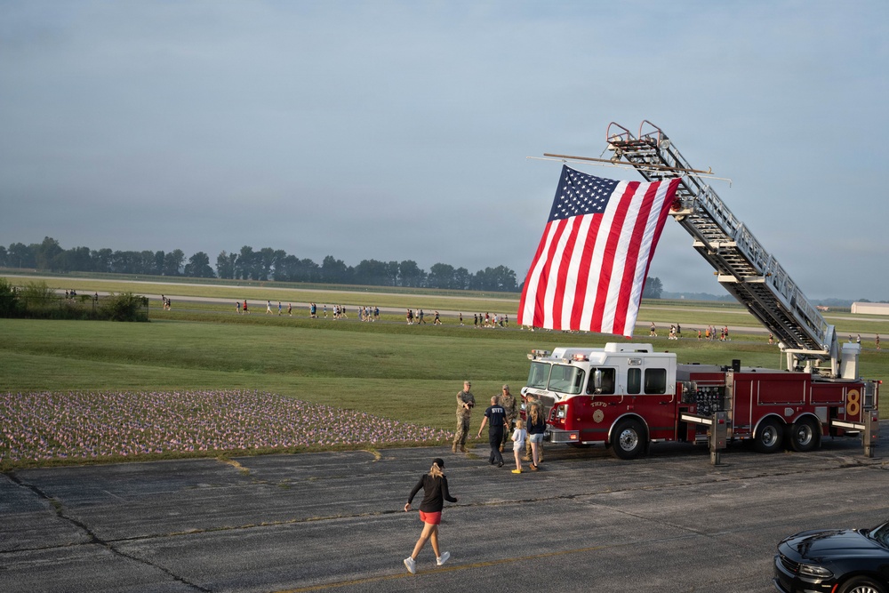 A large American flag is displayed from the extended aerial ladder of a red fire truck numbered '8' at what appears to be a military airfield or base. A field of small American flags is planted in the grass nearby, suggesting a memorial or patriotic commemoration event. A group of people can be seen walking along the airfield in the background, likely participating in a organized run or march.