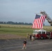 A large American flag is displayed from the extended aerial ladder of a red fire truck numbered '8' at what appears to be a military airfield or base. A field of small American flags is planted in the grass nearby, suggesting a memorial or patriotic commemoration event. A group of people can be seen walking along the airfield in the background, likely participating in a organized run or march.