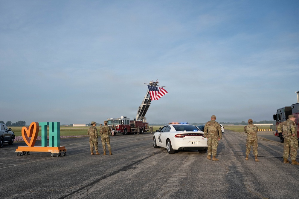 A military or official ceremony is taking place on an airport tarmac or airfield. A fire truck with an extended aerial ladder displays a large American flag, while military personnel in camouflage uniforms stand at attention alongside a white police/law enforcement vehicle with active emergency lights. A colorful 'TH' (Terre Haute) city sign is visible on the left side of the scene.