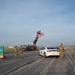 A military or official ceremony is taking place on an airport tarmac or airfield. A fire truck with an extended aerial ladder displays a large American flag, while military personnel in camouflage uniforms stand at attention alongside a white police/law enforcement vehicle with active emergency lights. A colorful 'TH' (Terre Haute) city sign is visible on the left side of the scene.