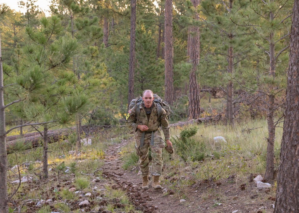 A male soldier in full camouflage uniform and tactical gear is rucking (marching with a loaded backpack) along a dirt trail through a pine forest. He carries a large military rucksack and appears to be mid-stride, looking directly at the camera. The setting suggests a training exercise or field operation in a mountainous or high-elevation forested environment.