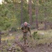 A male soldier in full camouflage uniform and tactical gear is rucking (marching with a loaded backpack) along a dirt trail through a pine forest. He carries a large military rucksack and appears to be mid-stride, looking directly at the camera. The setting suggests a training exercise or field operation in a mountainous or high-elevation forested environment.