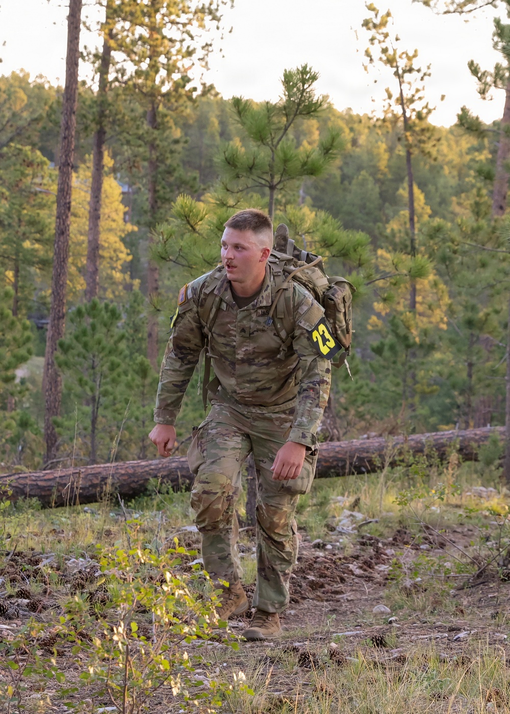 A soldier in US Army OCP (Operational Camouflage Pattern) uniform is conducting a ruck march through a pine forest terrain. The individual, wearing competitor number 23, is carrying a military rucksack and appears to be participating in a timed competition or training event. The setting appears to be a wooded hillside during early morning or late afternoon light.