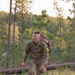 A soldier in US Army OCP (Operational Camouflage Pattern) uniform is conducting a ruck march through a pine forest terrain. The individual, wearing competitor number 23, is carrying a military rucksack and appears to be participating in a timed competition or training event. The setting appears to be a wooded hillside during early morning or late afternoon light.