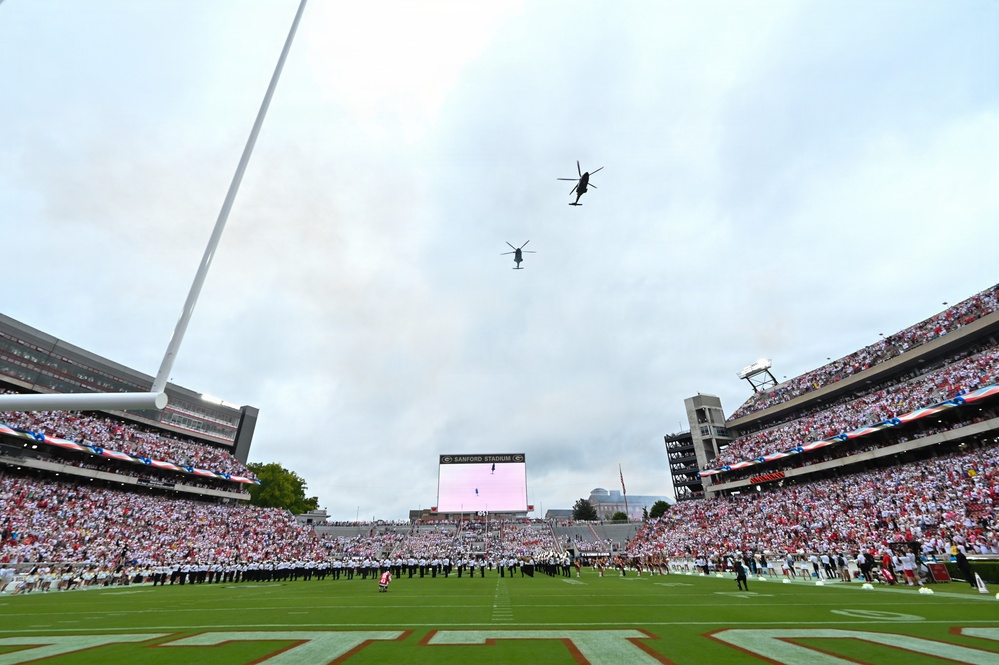 Two military helicopters perform a flyover above Sanford Stadium during a pre-game ceremony, likely at the University of Georgia. The stadium is packed with tens of thousands of fans dressed predominantly in red and white, while a marching band is assembled on the field below. Patriotic decorations are visible on the stadium facades, suggesting a military appreciation or opening ceremony event.