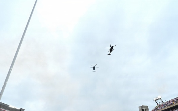 Two military helicopters perform a flyover above Sanford Stadium during a pre-game ceremony, likely at the University of Georgia. The stadium is packed with tens of thousands of fans dressed predominantly in red and white, while a marching band is assembled on the field below. Patriotic decorations are visible on the stadium facades, suggesting a military appreciation or opening ceremony event.