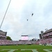 Two military helicopters perform a flyover above Sanford Stadium during a pre-game ceremony, likely at the University of Georgia. The stadium is packed with tens of thousands of fans dressed predominantly in red and white, while a marching band is assembled on the field below. Patriotic decorations are visible on the stadium facades, suggesting a military appreciation or opening ceremony event.