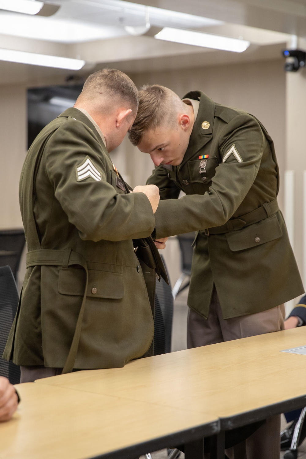 Two military personnel in olive green dress uniforms are leaning over a table in what appears to be a conference or classroom setting. The soldier on the right, wearing a Private First Class rank insignia and service ribbons, appears to be pinning or adjusting something on the uniform of the soldier on the left, who wears Sergeant chevrons. This appears to be a promotion or award ceremony.