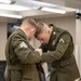 Two military personnel in olive green dress uniforms are leaning over a table in what appears to be a conference or classroom setting. The soldier on the right, wearing a Private First Class rank insignia and service ribbons, appears to be pinning or adjusting something on the uniform of the soldier on the left, who wears Sergeant chevrons. This appears to be a promotion or award ceremony.