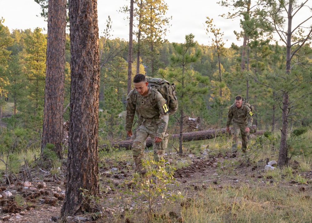 Two soldiers in military camouflage uniforms are conducting a ruck march through a pine forest, carrying large military rucksacks. Both wear numbered bibs (visible numbers 12 and 8), indicating this is a competitive military event or assessment. The setting appears to be a training area with sparse undergrowth, pine cones on the ground, and tall pine trees.