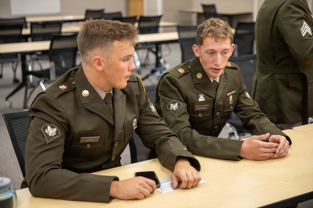 Two young soldiers in olive green dress uniforms are seated at a table in what appears to be a classroom or briefing room, engaged in conversation. Their name tags are partially visible, with the right soldier's tag reading 'MANHEMERT'. A third uniformed individual with sergeant stripes is partially visible in the background.