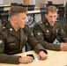 Two young soldiers in olive green dress uniforms are seated at a table in what appears to be a classroom or briefing room, engaged in conversation. Their name tags are partially visible, with the right soldier's tag reading 'MANHEMERT'. A third uniformed individual with sergeant stripes is partially visible in the background.