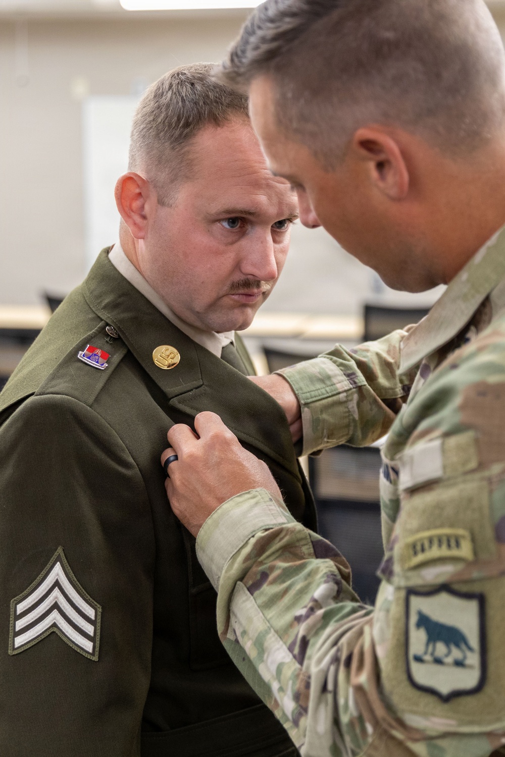 A soldier in modern U.S. Army OCP (Operational Camouflage Pattern) uniform is pinning something onto the collar or lapel of another soldier wearing an older-style olive drab dress uniform. The soldier receiving the pin holds the rank of Sergeant, visible by the chevrons on his sleeve, and wears a unit crest and brass insignia on his uniform. This appears to be a military promotion or award ceremony taking place in an office or administrative environment.