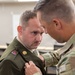 A soldier in modern U.S. Army OCP (Operational Camouflage Pattern) uniform is pinning something onto the collar or lapel of another soldier wearing an older-style olive drab dress uniform. The soldier receiving the pin holds the rank of Sergeant, visible by the chevrons on his sleeve, and wears a unit crest and brass insignia on his uniform. This appears to be a military promotion or award ceremony taking place in an office or administrative environment.