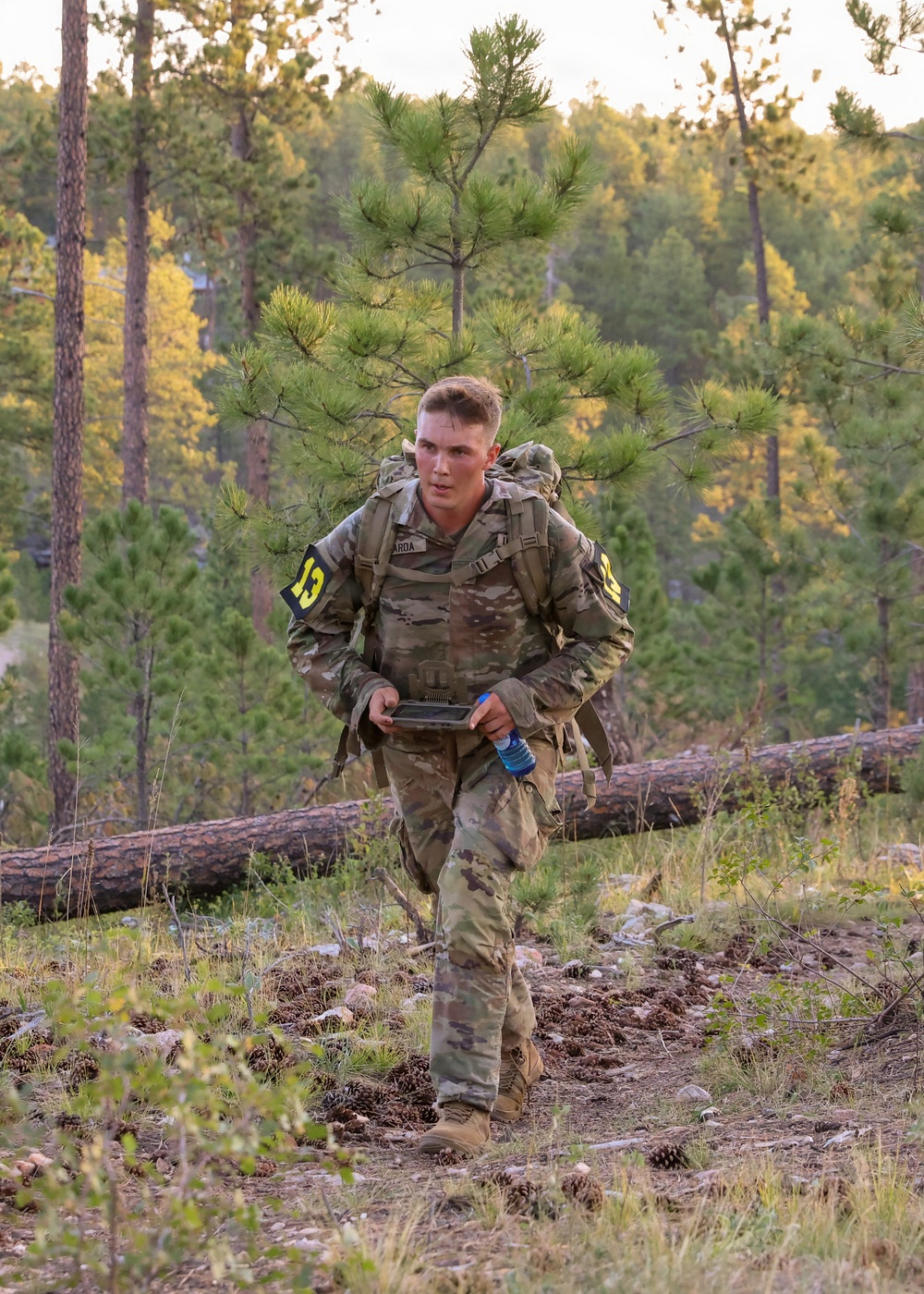 A young soldier in US Army OCP (Operational Camouflage Pattern) uniform is conducting a ruck march or land navigation exercise through a pine forest terrain. He is carrying a large rucksack on his back and holding what appears to be a compass or map case along with a water bottle. Yellow numbered patches on his shoulders suggest this is a training or competition event.