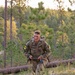 A young soldier in US Army OCP (Operational Camouflage Pattern) uniform is conducting a ruck march or land navigation exercise through a pine forest terrain. He is carrying a large rucksack on his back and holding what appears to be a compass or map case along with a water bottle. Yellow numbered patches on his shoulders suggest this is a training or competition event.