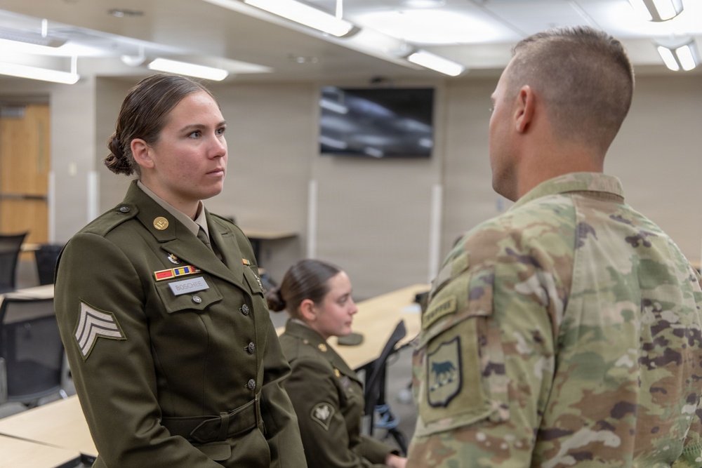 A U.S. Army female soldier in Army Service Uniform (ASU) is engaged in a face-to-face conversation with a male soldier wearing Operational Camouflage Pattern (OCP) uniform. The female soldier displays the rank of Sergeant (E-5) and several service ribbons on her dress uniform. The setting appears to be an indoor military classroom or conference room.