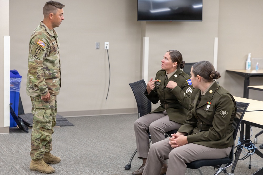 Three U.S. Army personnel are engaged in a conversation in what appears to be a modern briefing or classroom facility. One soldier in Operational Camouflage Pattern (OCP) uniform stands while two female soldiers in Army Green Service Uniforms (AGSU) are seated. The seated soldier appears to be actively speaking and gesturing, possibly pointing to insignia on her uniform.
