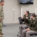 Three U.S. Army personnel are engaged in a conversation in what appears to be a modern briefing or classroom facility. One soldier in Operational Camouflage Pattern (OCP) uniform stands while two female soldiers in Army Green Service Uniforms (AGSU) are seated. The seated soldier appears to be actively speaking and gesturing, possibly pointing to insignia on her uniform.