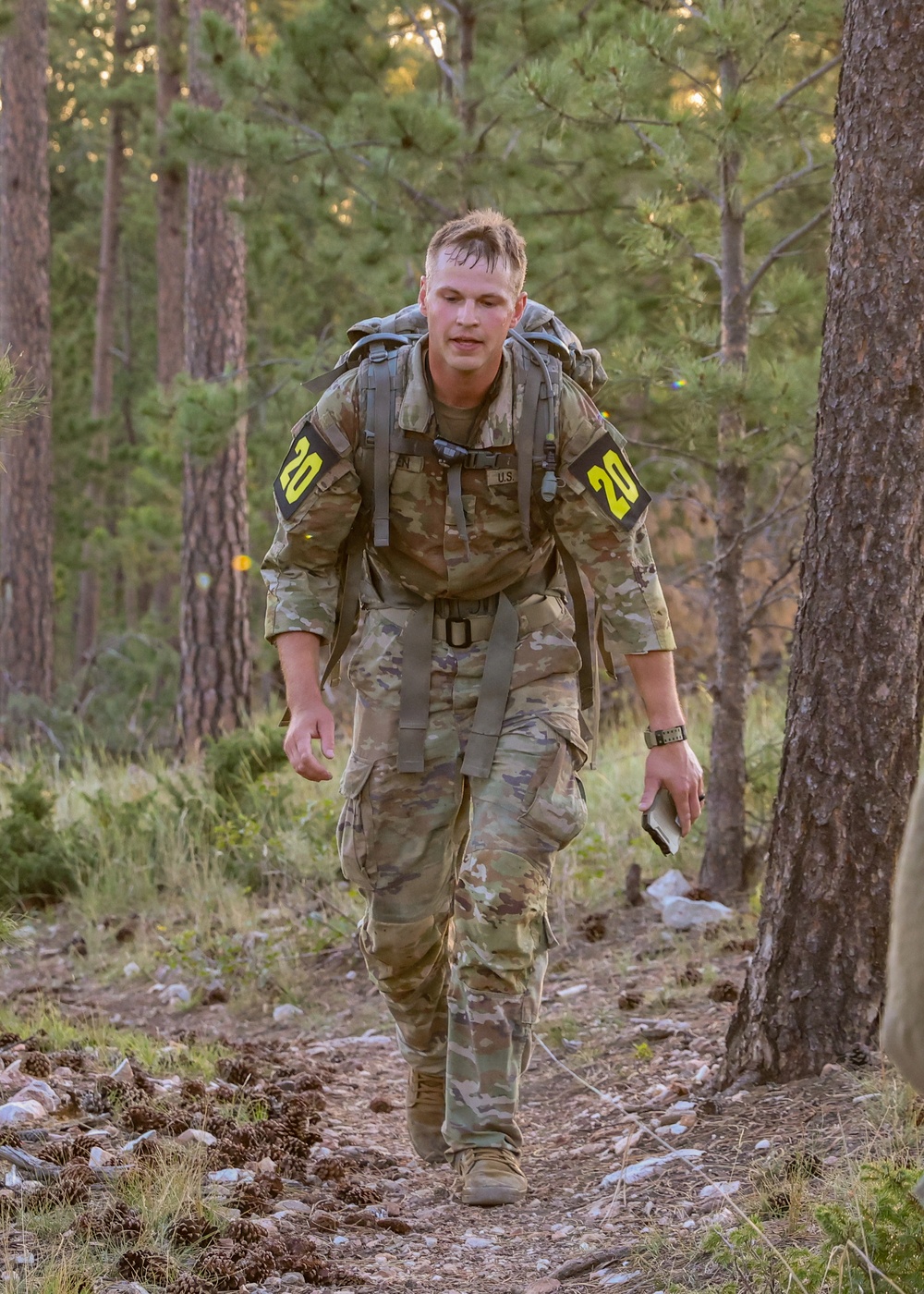 A U.S. Army soldier in OCP (Operational Camouflage Pattern) uniform is conducting a ruck march through a pine forest trail. The soldier wears competitor number '20' on both shoulders and carries a large military rucksack, suggesting participation in a timed military fitness competition or evaluation. The soldier appears physically exerted, with visible sweat and a forward-leaning posture.