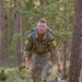 A U.S. Army soldier in OCP (Operational Camouflage Pattern) uniform is conducting a ruck march through a pine forest trail. The soldier wears competitor number '20' on both shoulders and carries a large military rucksack, suggesting participation in a timed military fitness competition or evaluation. The soldier appears physically exerted, with visible sweat and a forward-leaning posture.