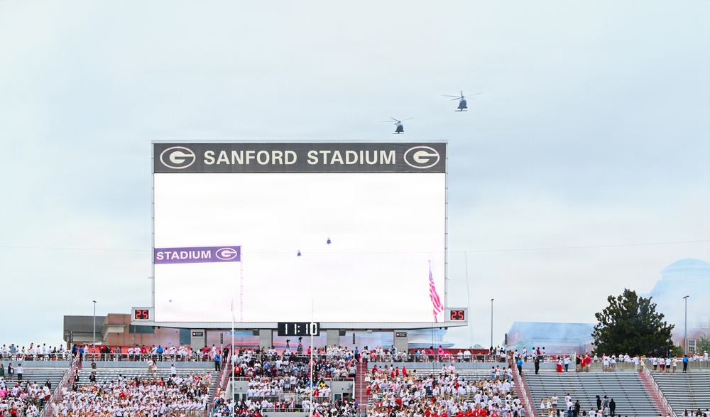The image shows Sanford Stadium, home of the University of Georgia Bulldogs, during a pre-game or ceremonial event. Two helicopters are flying over the stadium while crowds fill the stands, and an American flag is visible near the field. Smoke effects and the large video scoreboard displaying '11:10' suggest a special event or game-day ceremony.