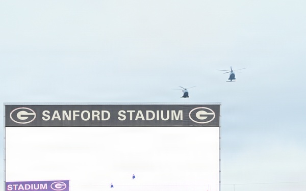 The image shows Sanford Stadium, home of the University of Georgia Bulldogs, during a pre-game or ceremonial event. Two helicopters are flying over the stadium while crowds fill the stands, and an American flag is visible near the field. Smoke effects and the large video scoreboard displaying '11:10' suggest a special event or game-day ceremony.