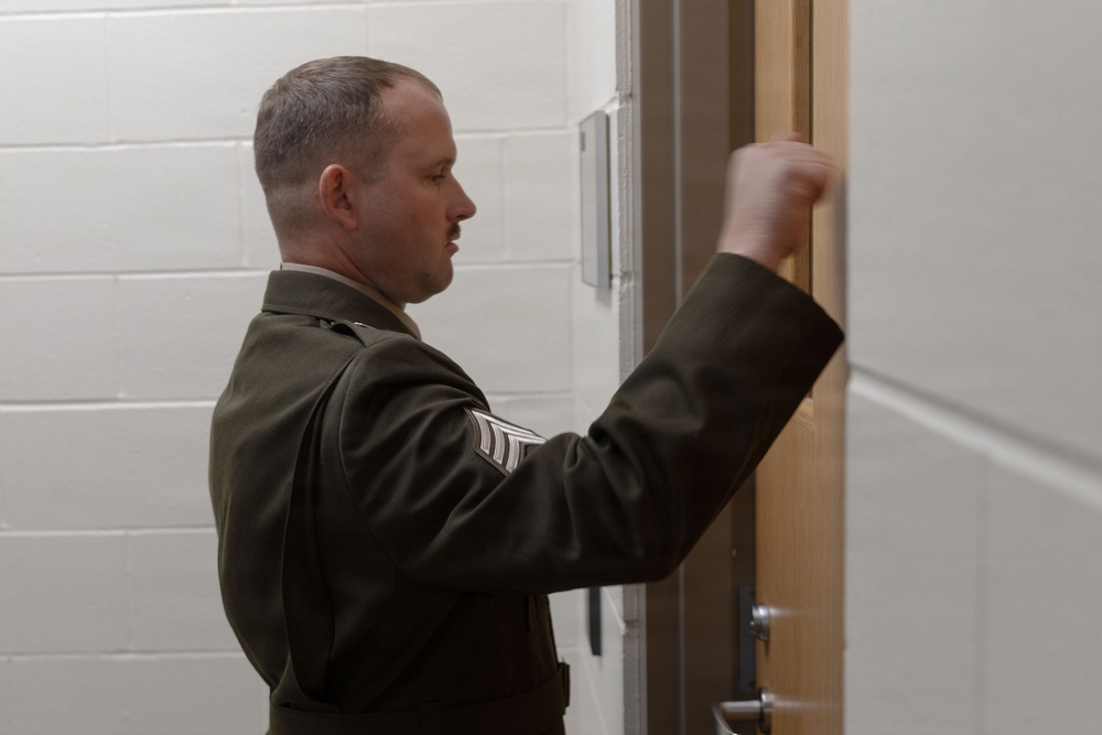 A male military service member in dress uniform is knocking on a door in what appears to be an institutional or administrative building. He is wearing an olive/dark green uniform with visible rank insignia on the sleeve, consistent with a U.S. Marine Corps dress uniform. His raised fist is captured mid-knock against a wooden door frame.