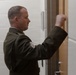 A male military service member in dress uniform is knocking on a door in what appears to be an institutional or administrative building. He is wearing an olive/dark green uniform with visible rank insignia on the sleeve, consistent with a U.S. Marine Corps dress uniform. His raised fist is captured mid-knock against a wooden door frame.