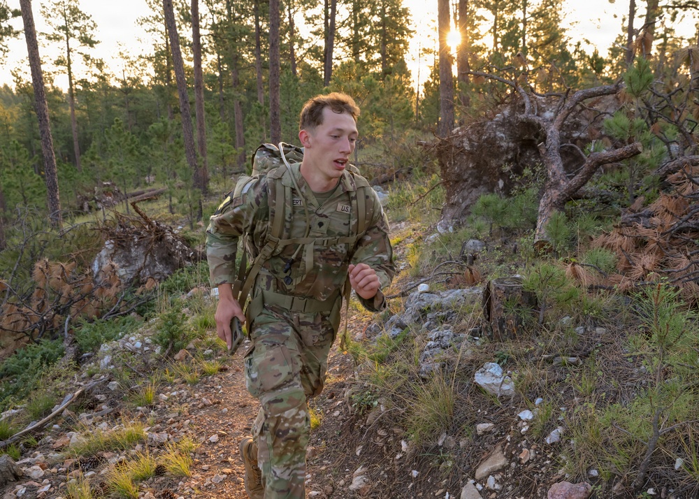 A U.S. Army soldier in full OCP (Operational Camouflage Pattern) uniform is conducting a ruck march or physical training exercise through a rocky, forested terrain. The soldier is carrying a military rucksack and appears to be moving at a fast pace along a narrow trail. The image is taken during golden hour, with sunlight filtering through pine trees in the background.