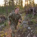 A U.S. Army soldier in full OCP (Operational Camouflage Pattern) uniform is conducting a ruck march or physical training exercise through a rocky, forested terrain. The soldier is carrying a military rucksack and appears to be moving at a fast pace along a narrow trail. The image is taken during golden hour, with sunlight filtering through pine trees in the background.