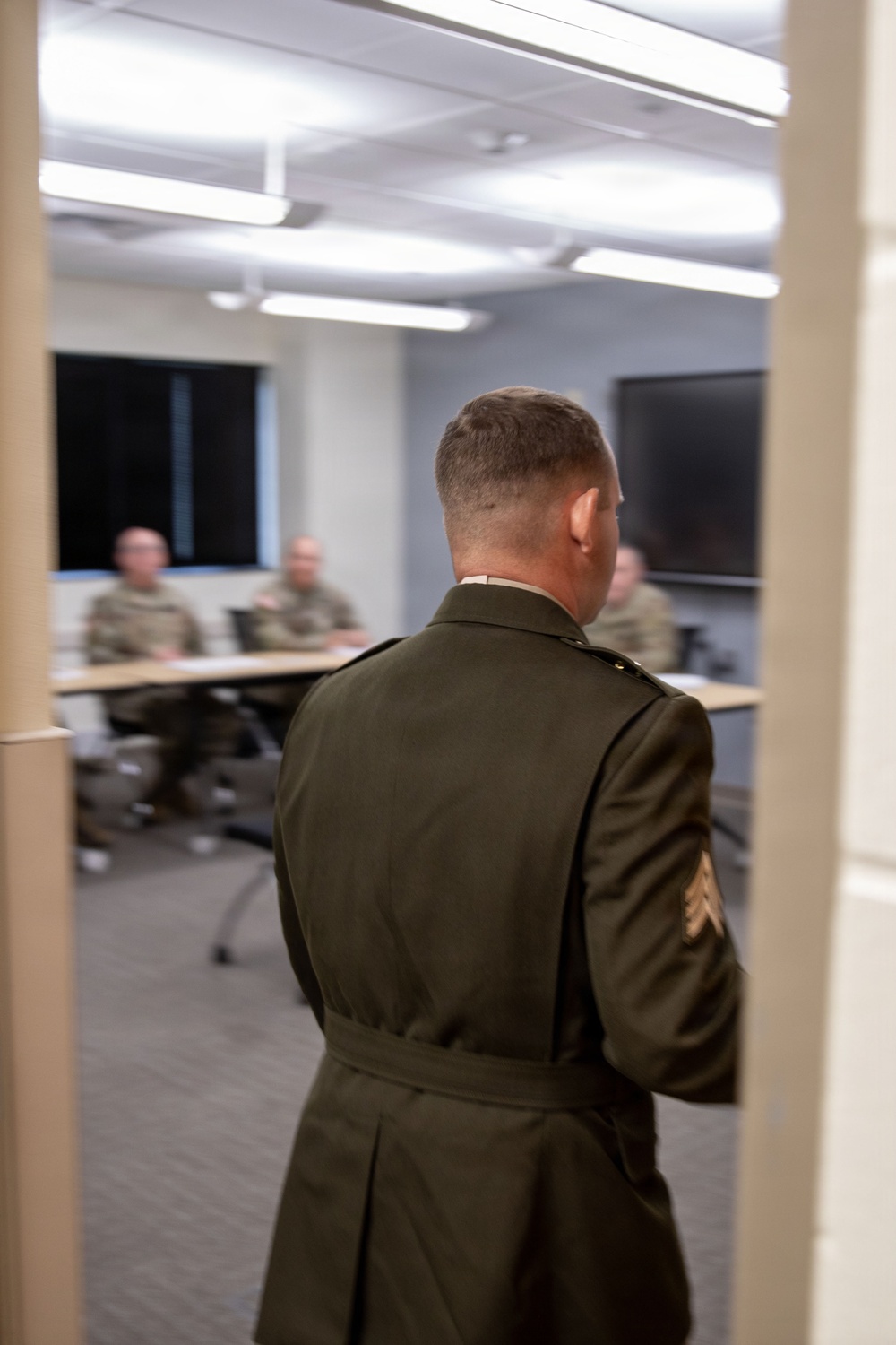A military personnel member in a dark olive dress uniform is viewed from behind, standing in a doorway facing a conference or briefing room. Inside the room, several individuals in military combat uniforms (ACU/OCP pattern) are seated at tables. The scene appears to depict a formal briefing or meeting setting.