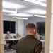 A military personnel member in a dark olive dress uniform is viewed from behind, standing in a doorway facing a conference or briefing room. Inside the room, several individuals in military combat uniforms (ACU/OCP pattern) are seated at tables. The scene appears to depict a formal briefing or meeting setting.