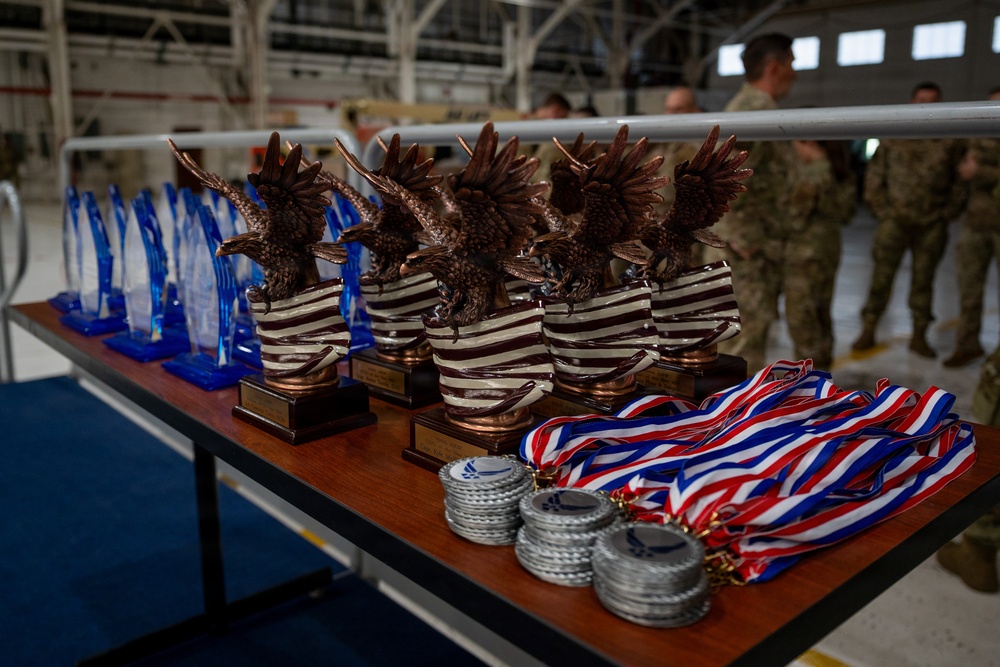 A display table in what appears to be a military hangar holds multiple awards arranged for a ceremony. The table features bronze eagle trophies on striped bases, blue acrylic trophies, and stacks of medals with red, white, and blue ribbons. Military personnel in camouflage uniforms are visible in the blurred background.