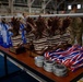 A display table in what appears to be a military hangar holds multiple awards arranged for a ceremony. The table features bronze eagle trophies on striped bases, blue acrylic trophies, and stacks of medals with red, white, and blue ribbons. Military personnel in camouflage uniforms are visible in the blurred background.