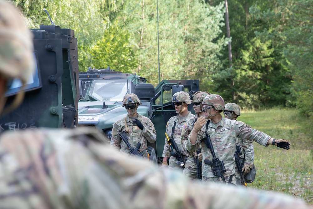 A group of U.S. Army soldiers in full combat gear are gathered near military vehicles in a wooded outdoor area, appearing to conduct a briefing or coordination discussion. The soldiers are wearing MultiCam (OCP) uniforms, helmets, protective eyewear, and carrying rifles. The setting suggests a field training exercise or operational deployment.