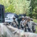 A group of U.S. Army soldiers in full combat gear are gathered near military vehicles in a wooded outdoor area, appearing to conduct a briefing or coordination discussion. The soldiers are wearing MultiCam (OCP) uniforms, helmets, protective eyewear, and carrying rifles. The setting suggests a field training exercise or operational deployment.