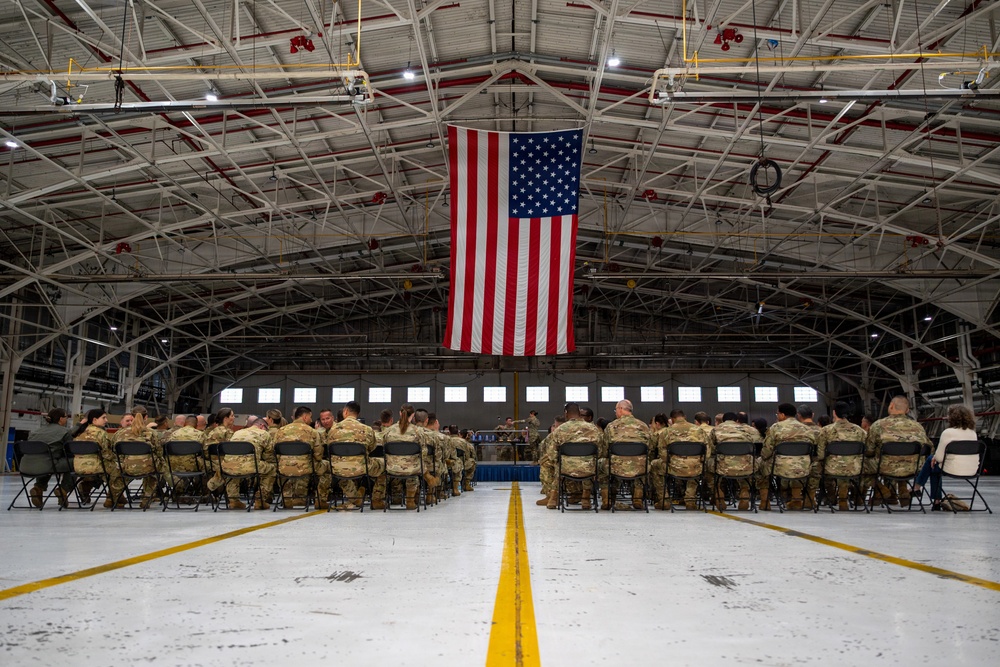 A military formation ceremony is taking place inside a large aircraft hangar, with uniformed personnel seated in rows of folding chairs facing a stage or podium area. A large American flag is prominently displayed hanging from the ceiling rafters at the center of the hangar. The setting and attire suggest an official military event such as a deployment ceremony, award ceremony, or unit formation.