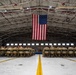 A military formation ceremony is taking place inside a large aircraft hangar, with uniformed personnel seated in rows of folding chairs facing a stage or podium area. A large American flag is prominently displayed hanging from the ceiling rafters at the center of the hangar. The setting and attire suggest an official military event such as a deployment ceremony, award ceremony, or unit formation.