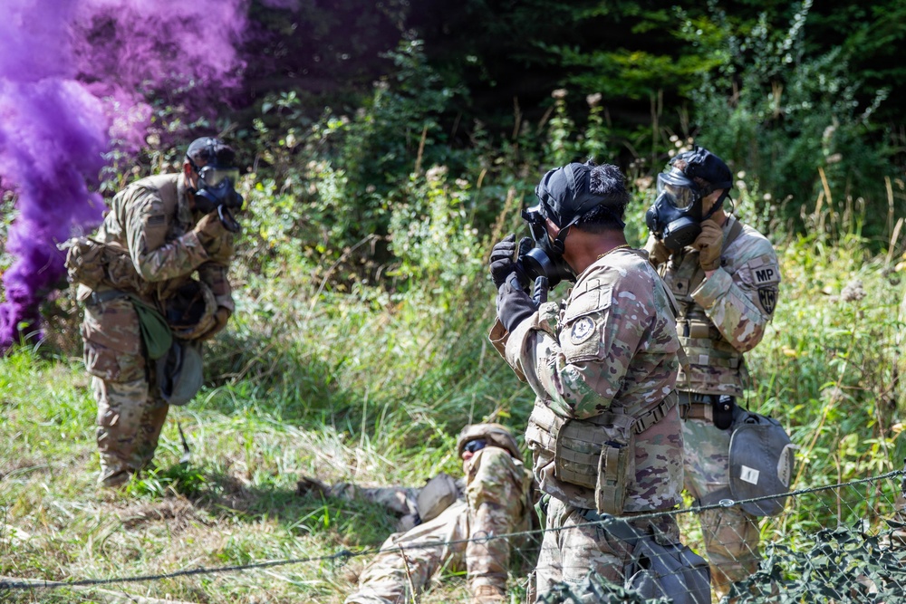 Military personnel in multicam/OCP uniforms are conducting a field training exercise involving colored smoke grenades. Multiple soldiers are wearing gas masks and tactical gear in an outdoor, vegetated environment. One soldier visible on the right has 'MP' (Military Police) markings on their vest.