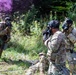 Military personnel in multicam/OCP uniforms are conducting a field training exercise involving colored smoke grenades. Multiple soldiers are wearing gas masks and tactical gear in an outdoor, vegetated environment. One soldier visible on the right has 'MP' (Military Police) markings on their vest.