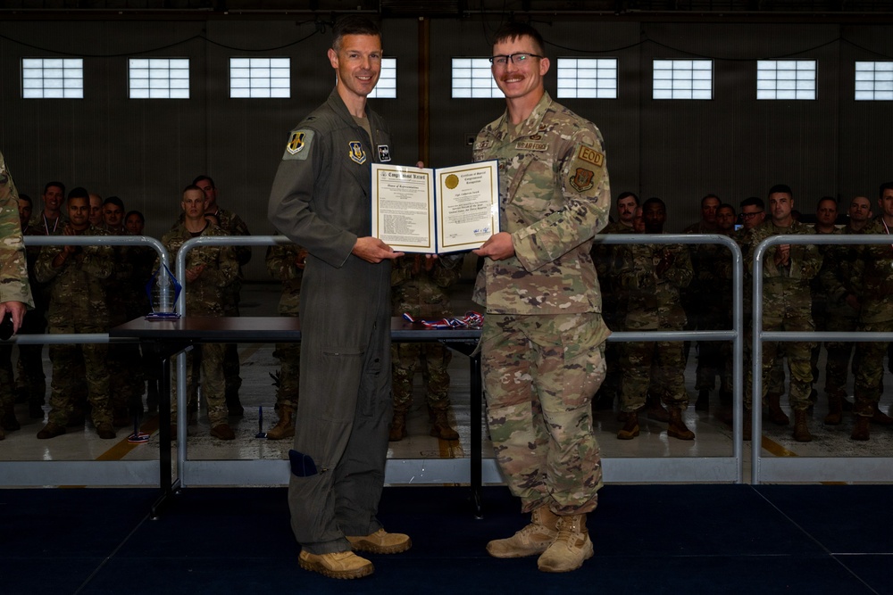 Two U.S. Air Force personnel are posing together holding a military commendation or award certificate during a formal ceremony inside an aircraft hangar. The individual on the left is wearing an olive green flight suit, while the person on the right is in OCP (Operational Camouflage Pattern) uniform with an EOD patch visible. A formation of uniformed military personnel is visible in the background observing the ceremony.