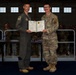 Two U.S. Air Force personnel are posing together holding a military commendation or award certificate during a formal ceremony inside an aircraft hangar. The individual on the left is wearing an olive green flight suit, while the person on the right is in OCP (Operational Camouflage Pattern) uniform with an EOD patch visible. A formation of uniformed military personnel is visible in the background observing the ceremony.
