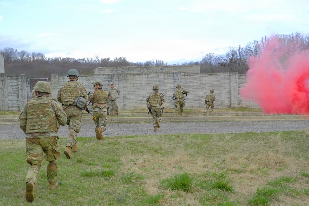 A group of military personnel in camouflage uniforms are running toward a concrete wall structure during what appears to be a training exercise. A red smoke grenade is deployed on the right side of the image, likely used as a signal or to simulate battlefield conditions. The scene takes place on a military training facility with sparse grass and leafless trees in the background, suggesting late autumn or early spring.
